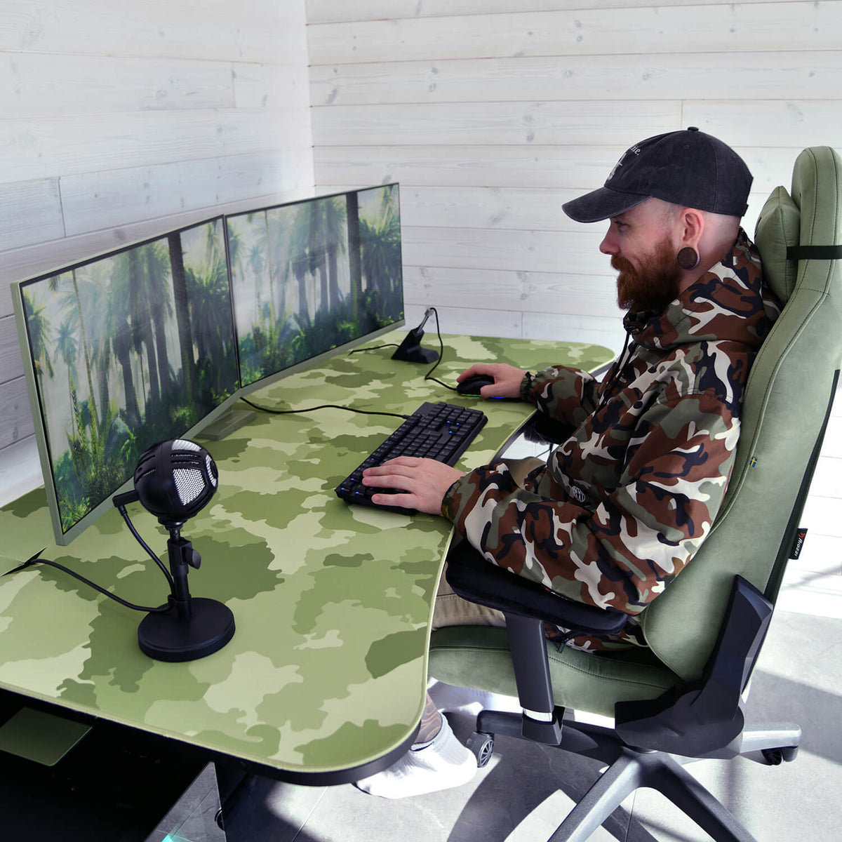 Man in camo jacket at curved desk with Arozzi Europe Arena Mousepad, triple monitors, and mic.