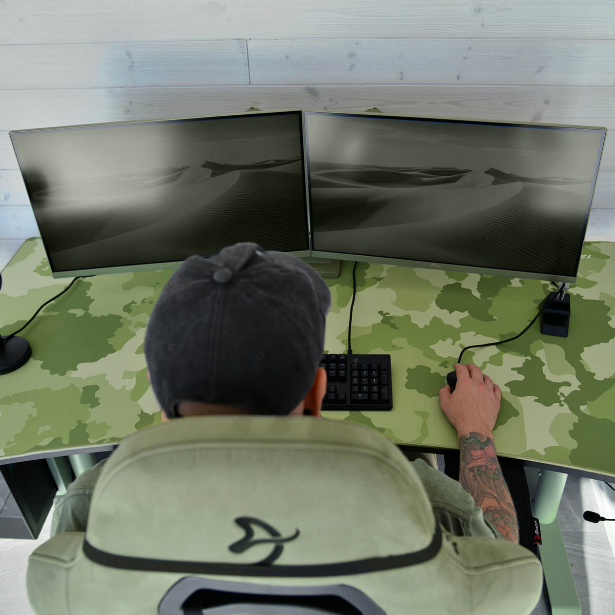 Person at dual monitors on a camo desk, using an Arozzi Europe Arena Mousepad, wearing a cap.
