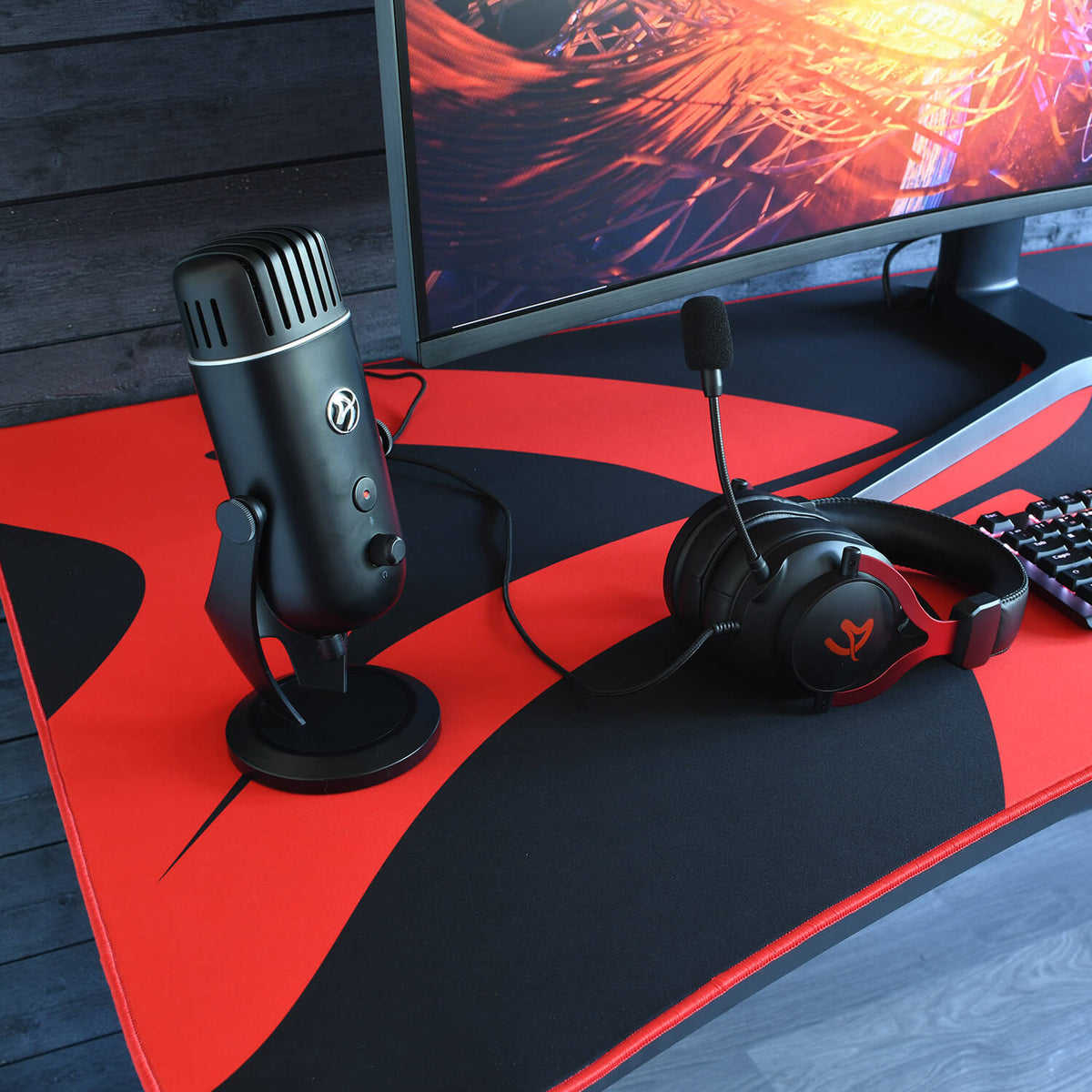 Black mic and headset on a gaming desk with an Arozzi Europe Arena Mousepad by a curved monitor.