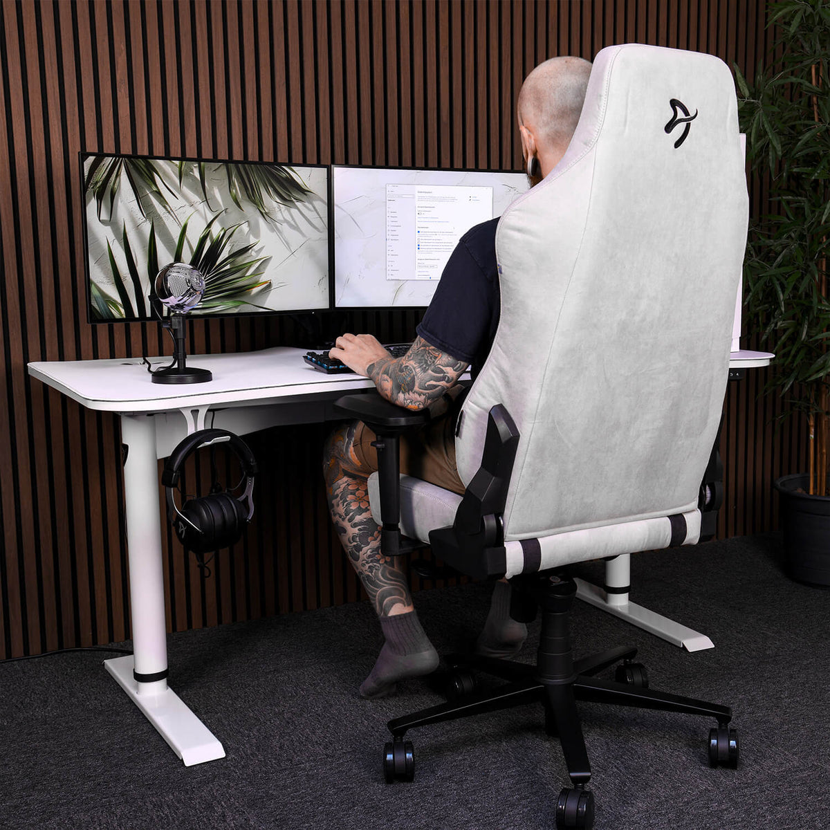 Tattooed person at desk with dual monitors, mic, and Arozzi Europe Headset Hanger.