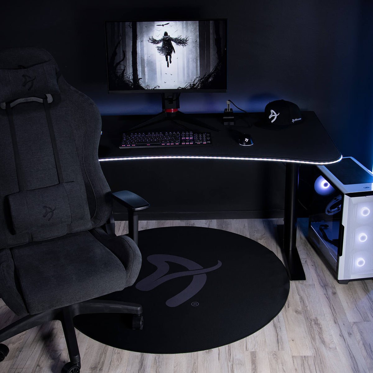 Dark gaming setup with curved desk, PC, chair, and Arozzi Zona Round Floor Pad on wood floor.