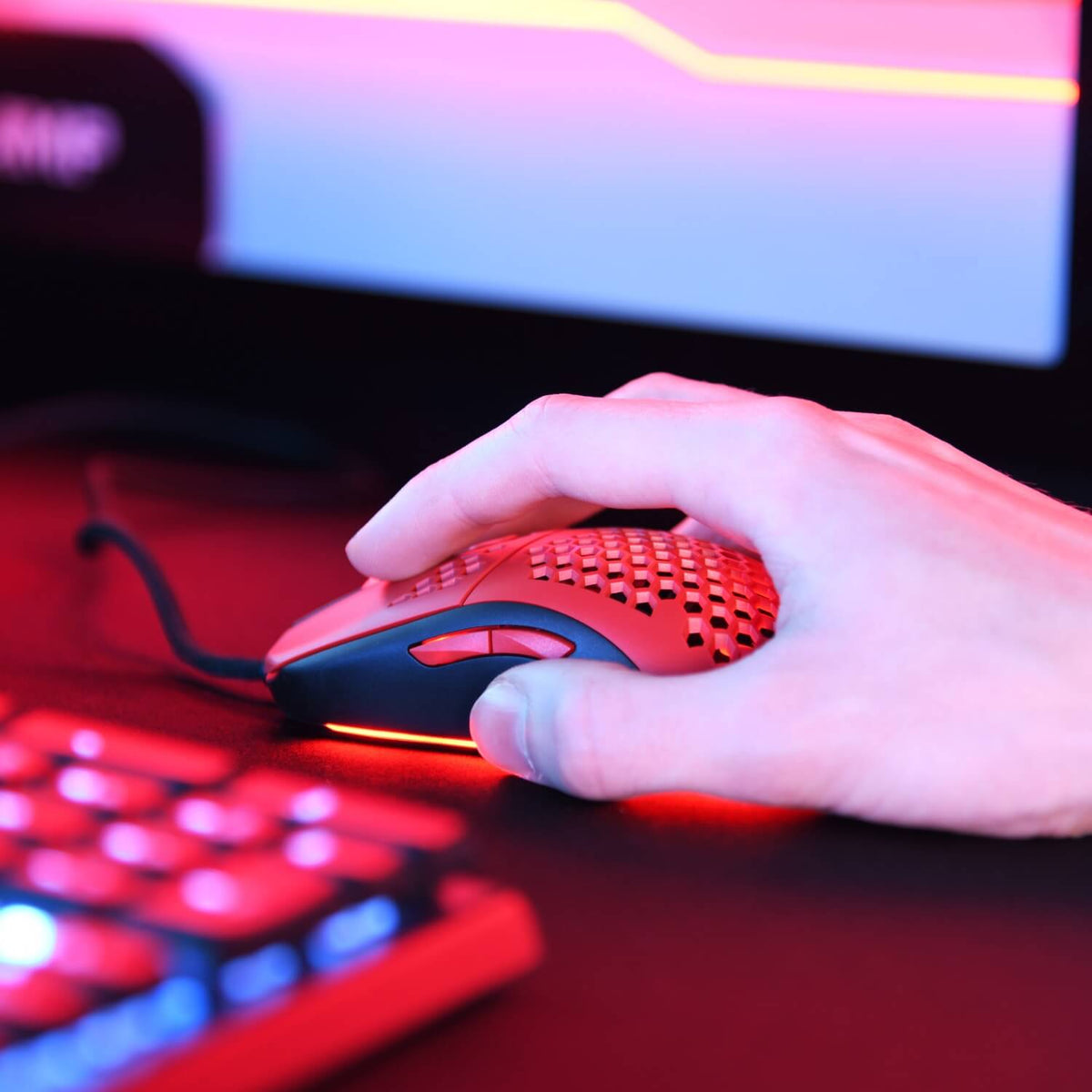 Hand on a Favo Ultra Light Gaming Mouse by Arozzi Europe, by a red-lit keyboard and monitor.
