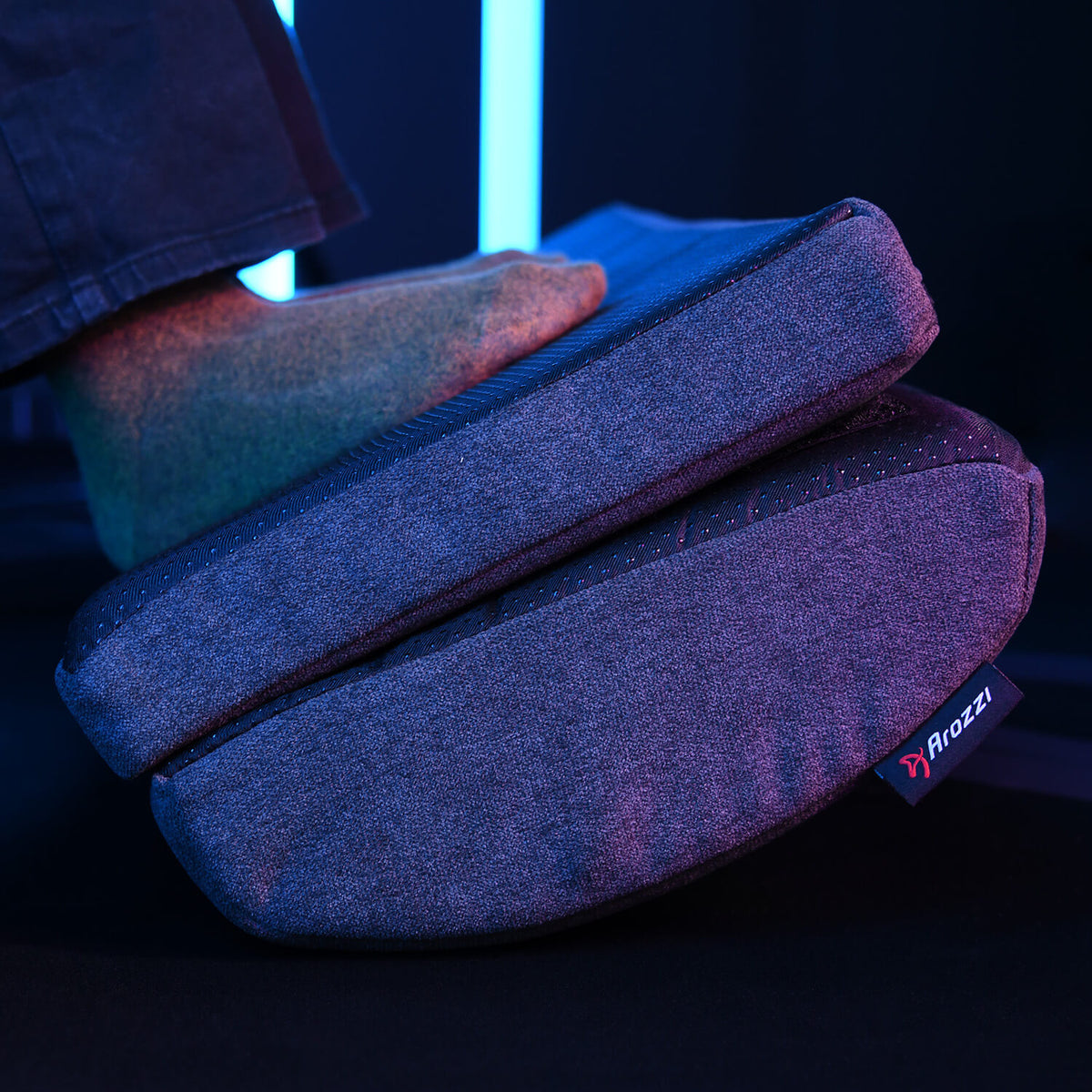 A foot rests on a gray Arozzi footrest cushion with blue lighting in the background.