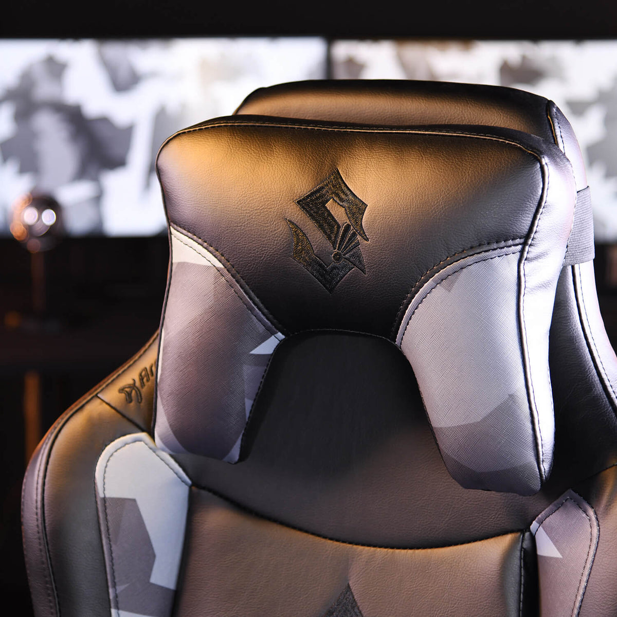 Black and gray gaming chair with headrest, logo, and blurred monitors in the background.