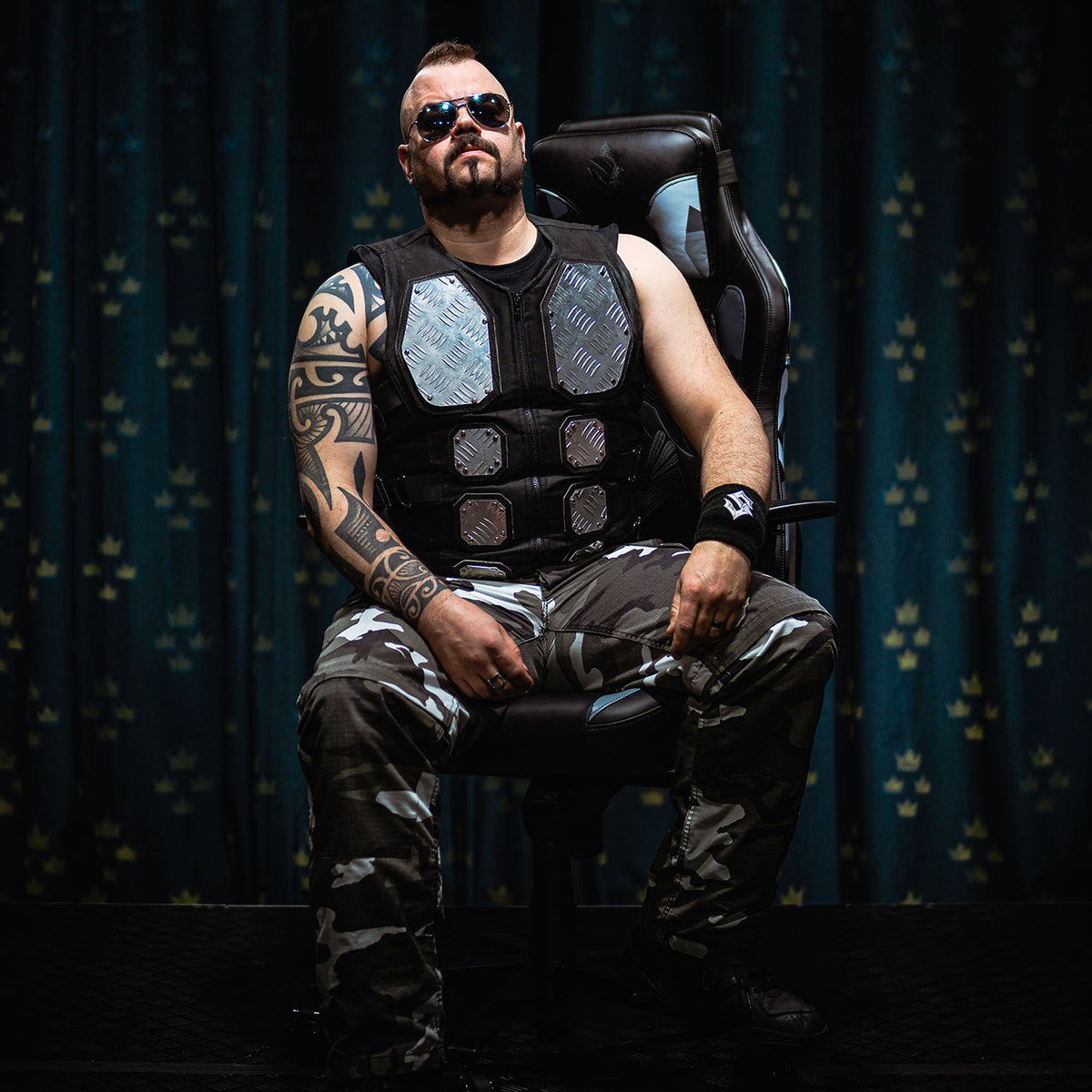 Man in camo pants, sunglasses & armor vest sits on Arozzi Vernazza Sabaton Edition gaming chair.