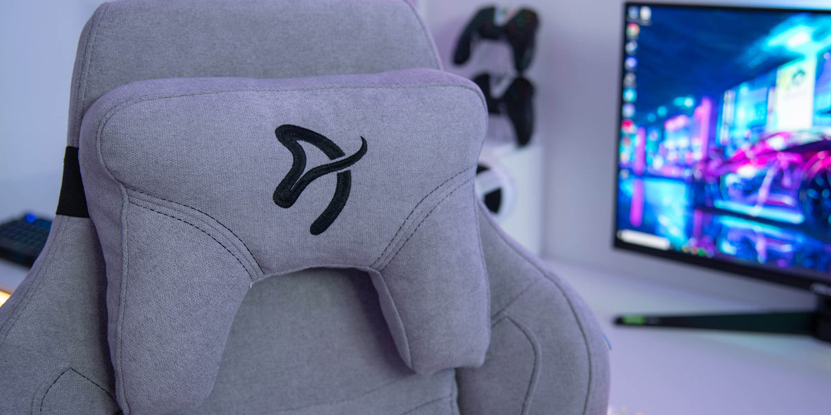 Gray gaming chair with black logo in front of a monitor and game controllers on the wall.