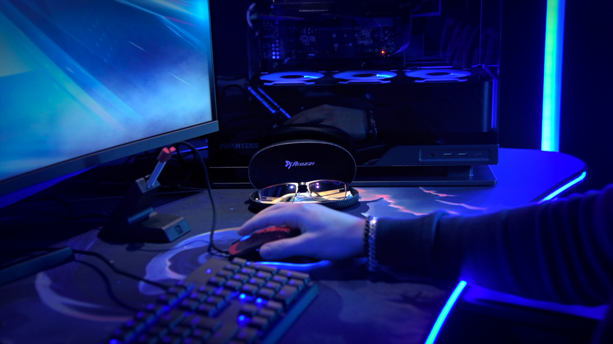 A person using a computer mouse next to a monitor, keyboard, and eyeglasses in blue light.