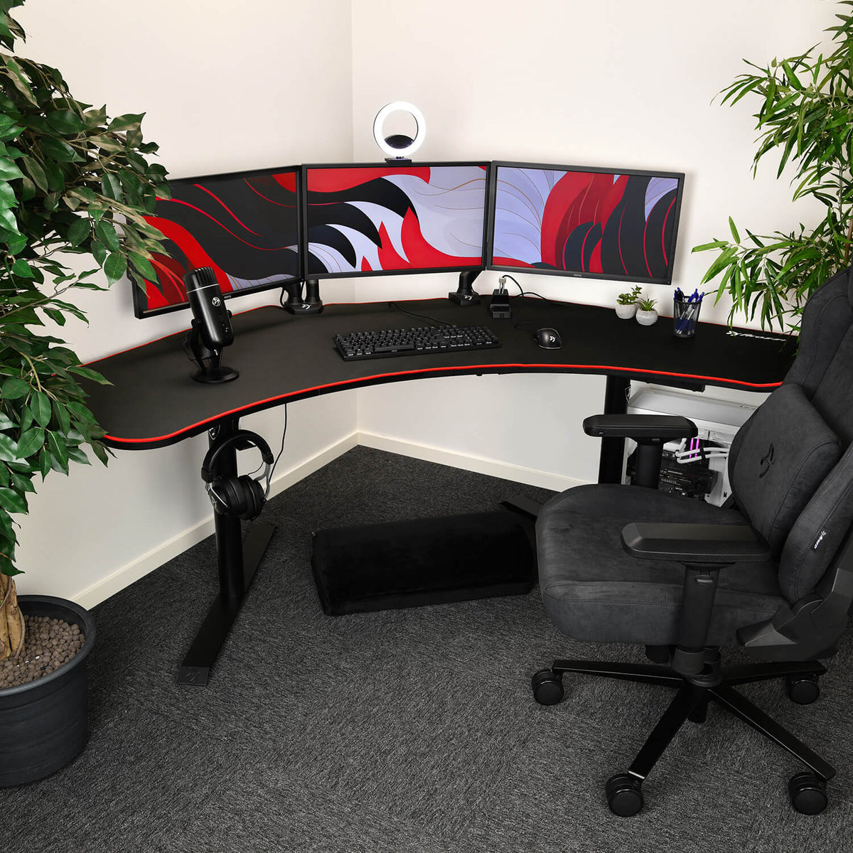 L-shaped desk setup with 3 monitors, mic, chair, plants, and Arozzi Arena Angelo Mousepad.