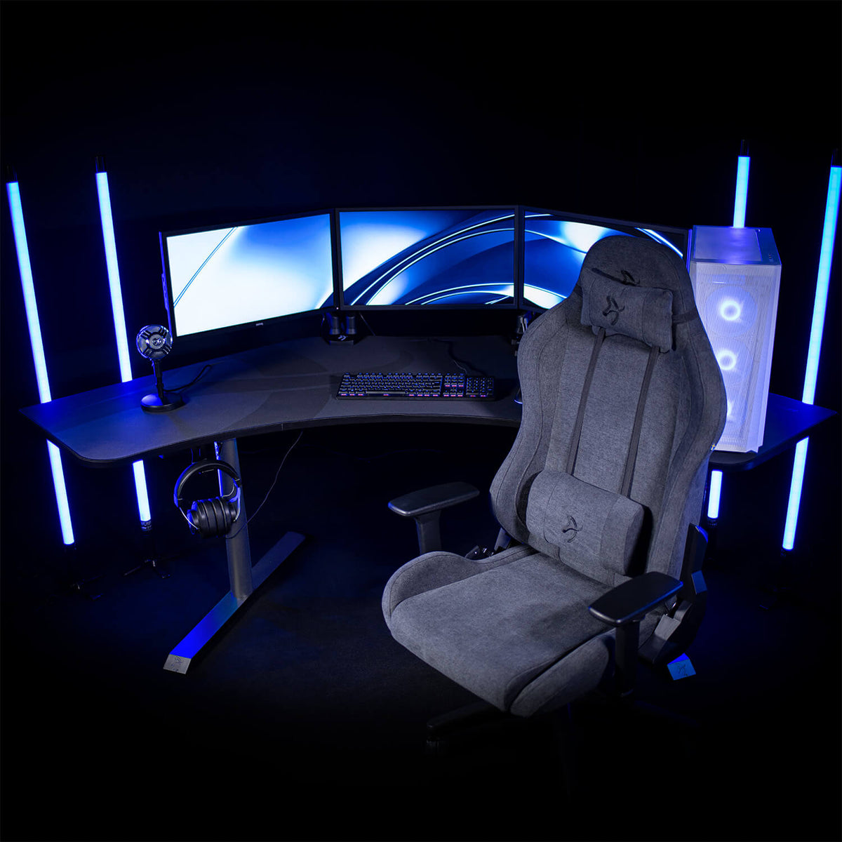 Arena Angelo corner desk by Arozzi Europe, triple monitors, LEDs, grey gaming chair in dark room.