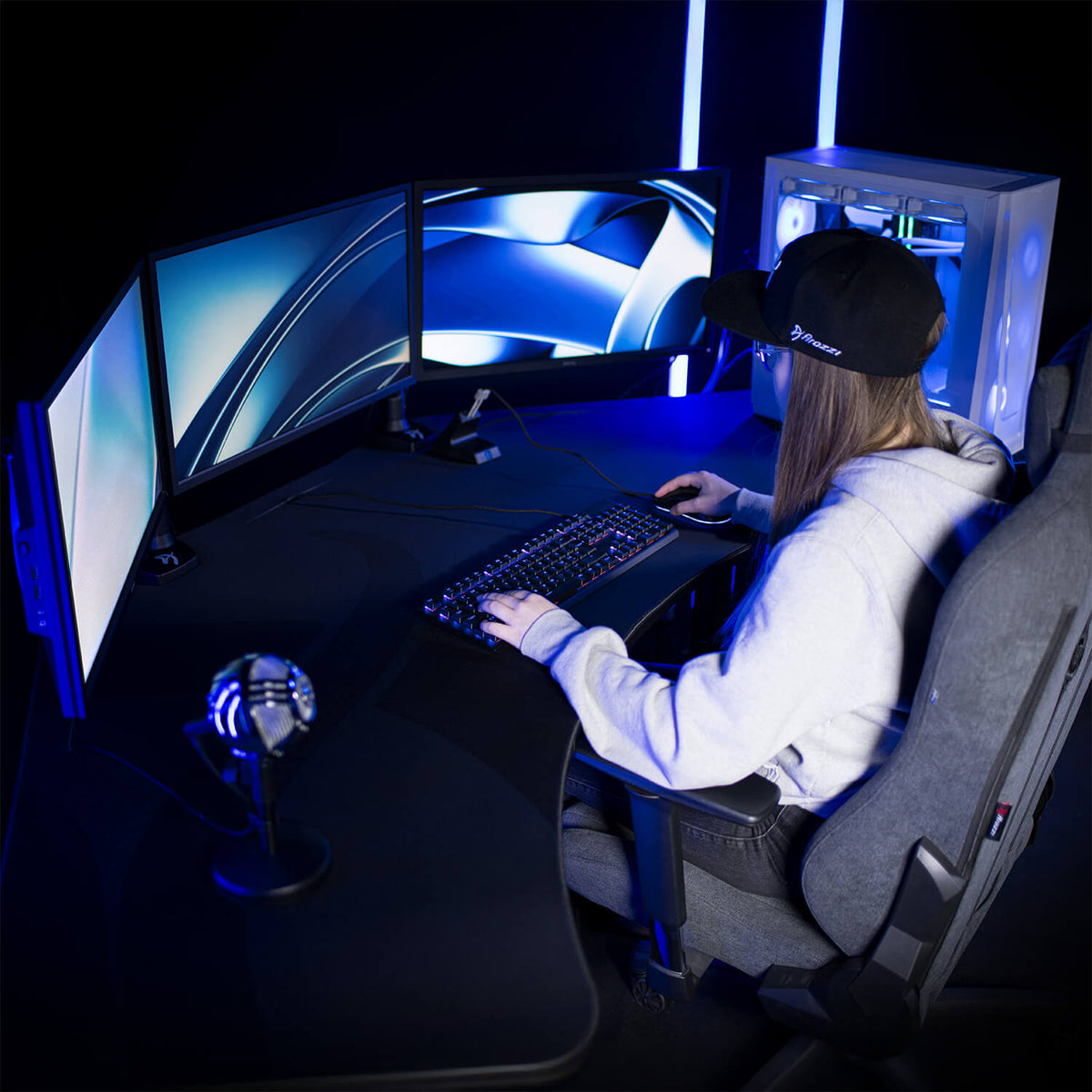 Gamer at Arena Angelo desk by Arozzi Europe with triple monitors, PC, mic, and blue lights.