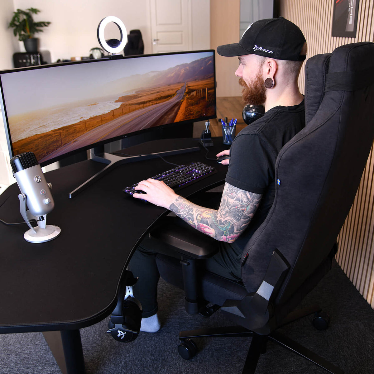 Tattooed man at Arena Angelo desk by Arozzi Europe with gaming setup, wide monitor, and mic.