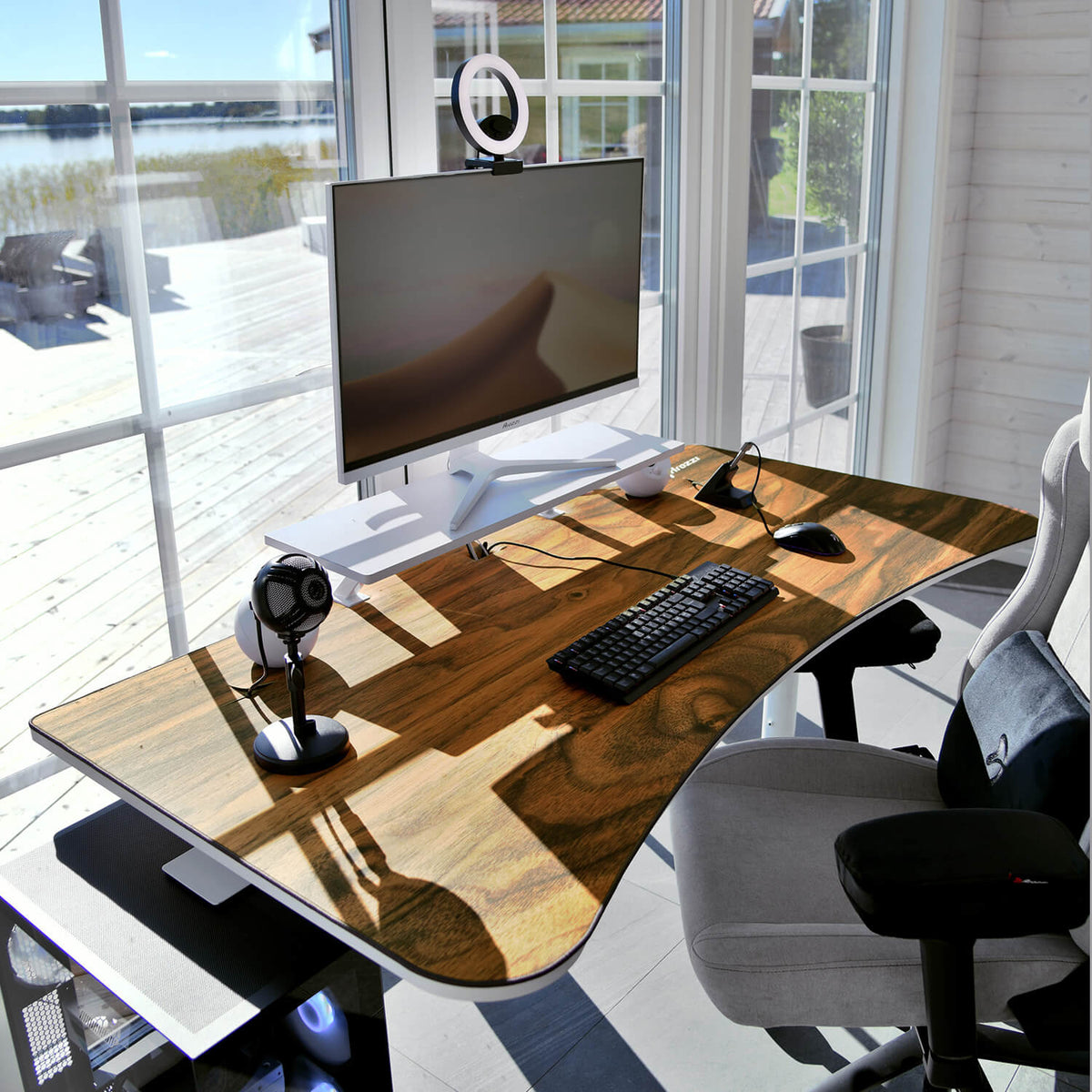 Modern desk with Arozzi Europe Arena Mousepad, monitor, keyboard, mic & headphones by sunny windows.