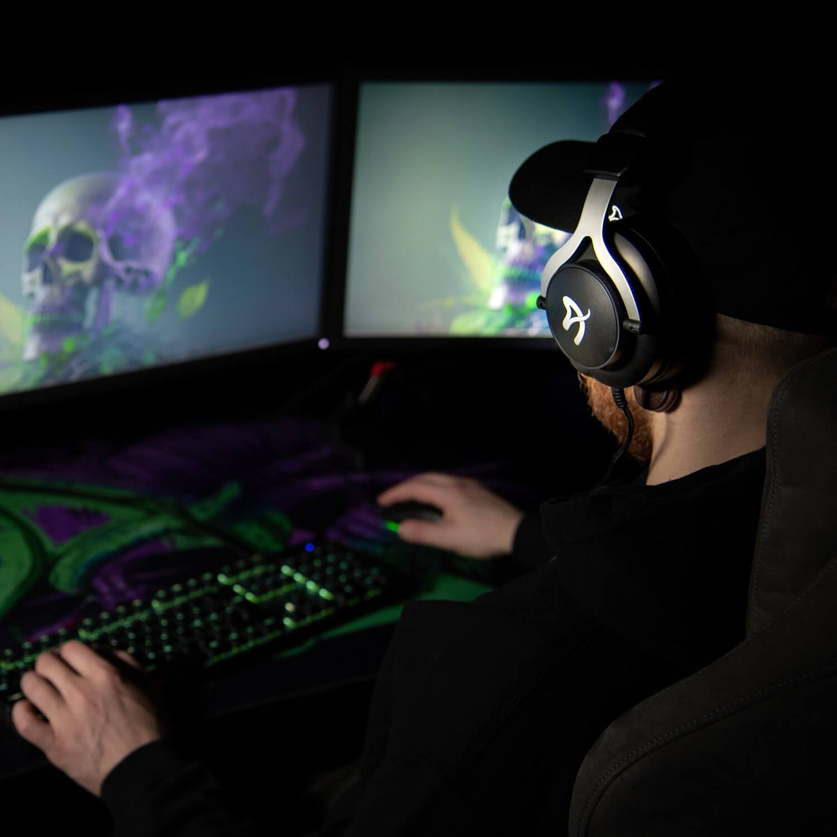 Gamer with Arozzi Europe Aria headset at dual monitors, skull graphic displayed onscreen.