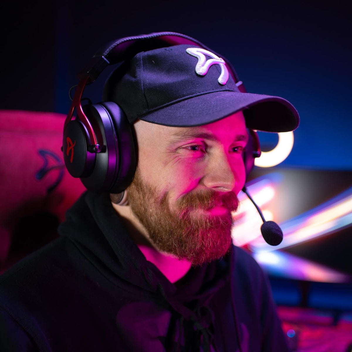 Bearded man in cap wears Arozzi Europe Aria Gaming Headset, pink-lit, at computer gaming setup.