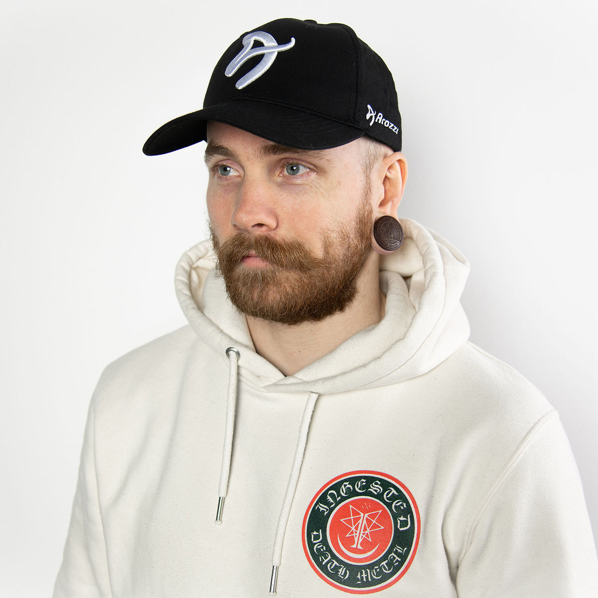 Man in white hoodie, Arozzi Europe Sport Cap, beard, ear gauges, white background.