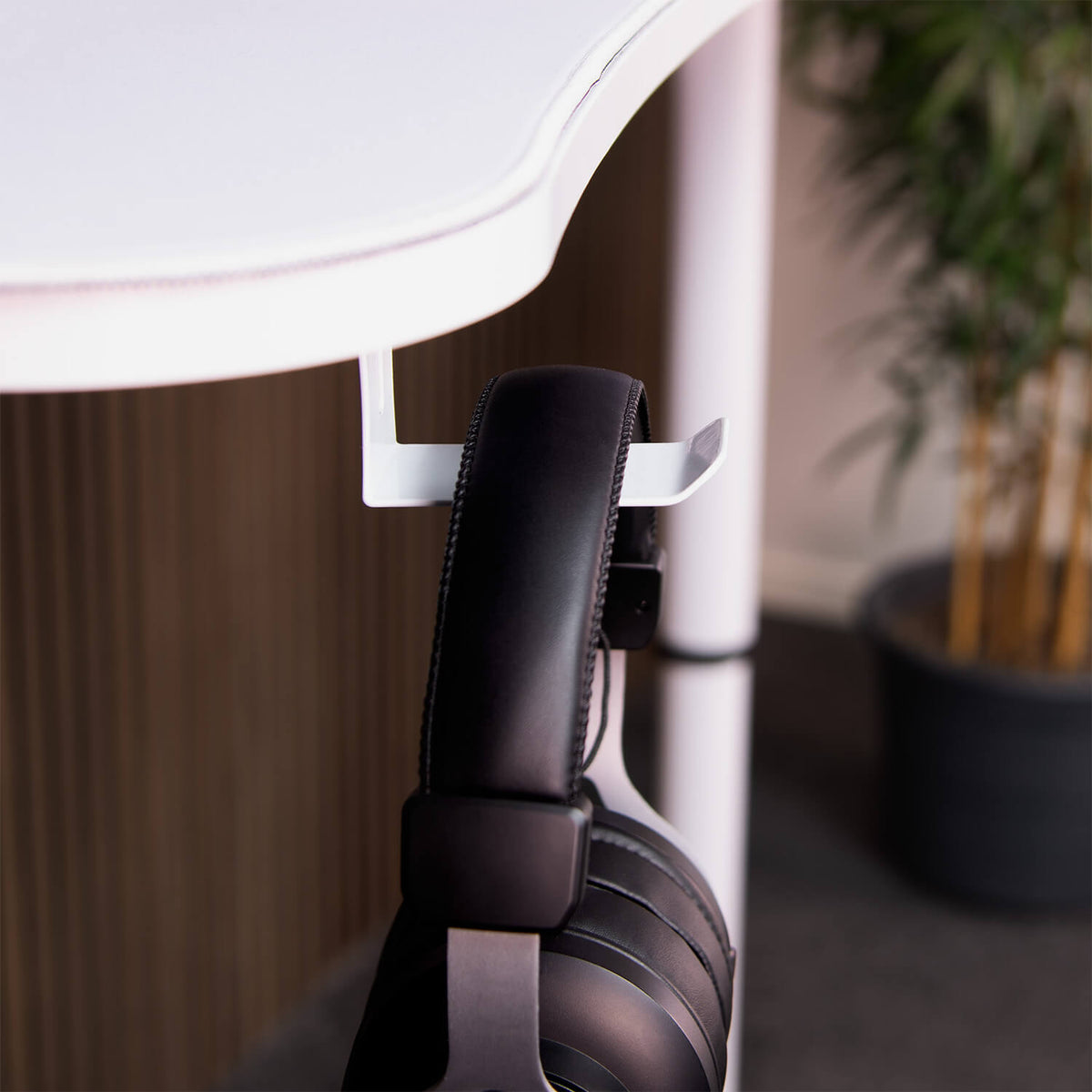 Arozzi Europe Headset Hanger holds black headphones beneath white desk; plant decorates background.