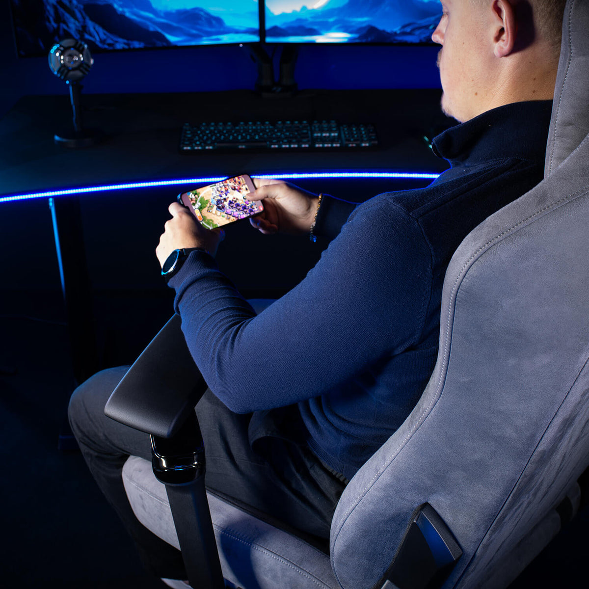 Gamer in Arozzi Europe’s chair with Infinity Armrests, playing on phone, monitors visible behind.