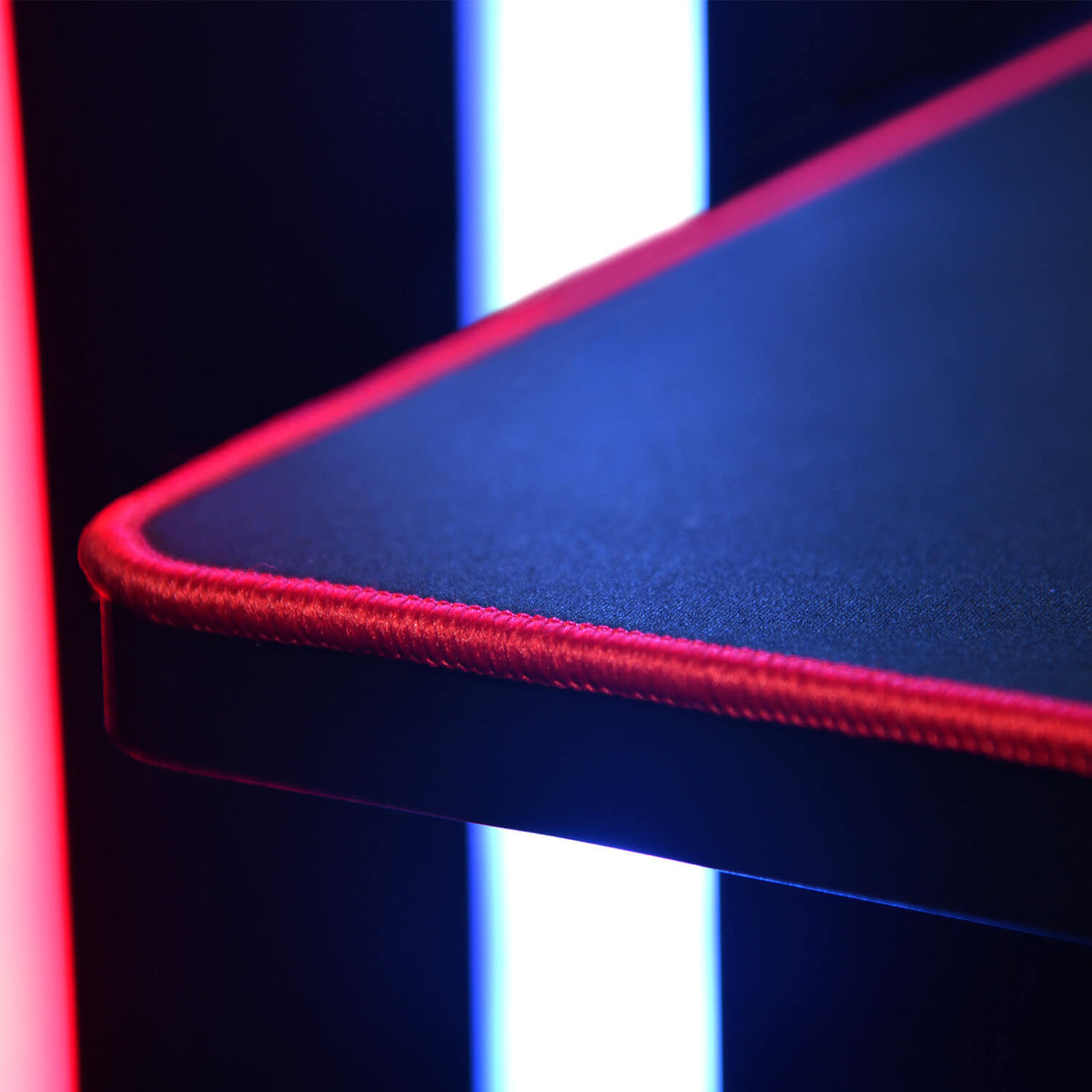 A close-up of a mouse pad corner with red stitching and neon lights in the background.