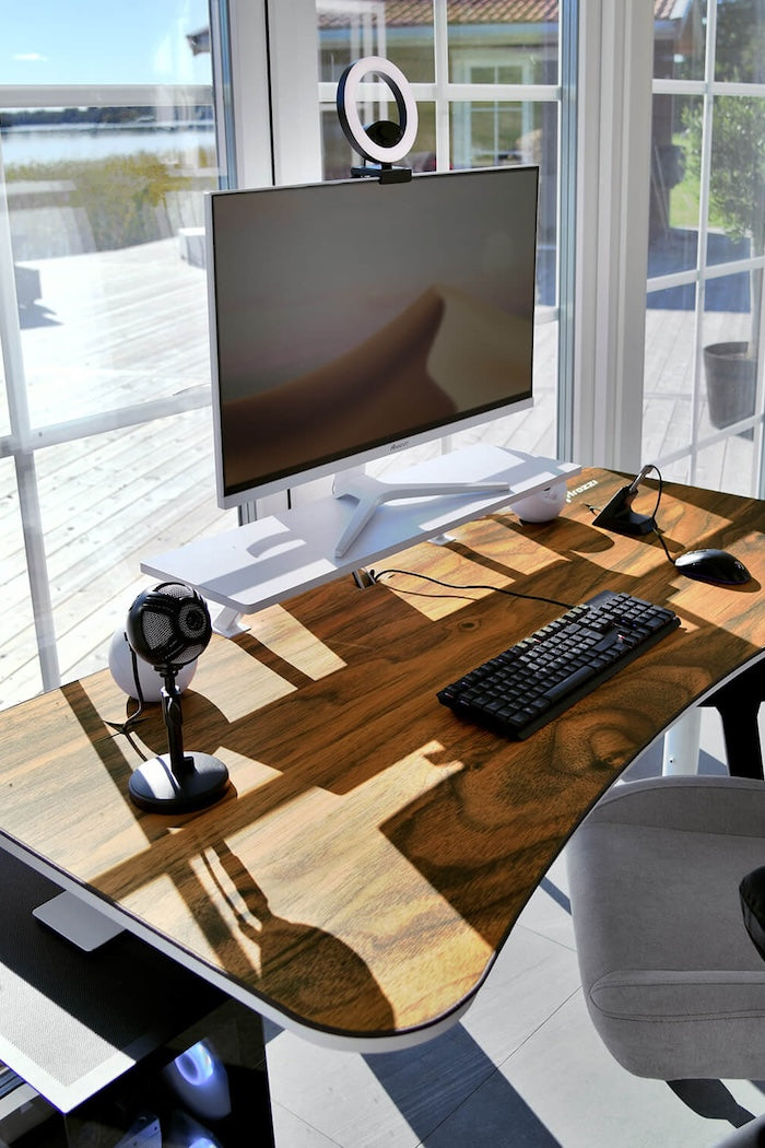 Modern desk setup with monitor, ring light, keyboard, and microphone by sunny windows.