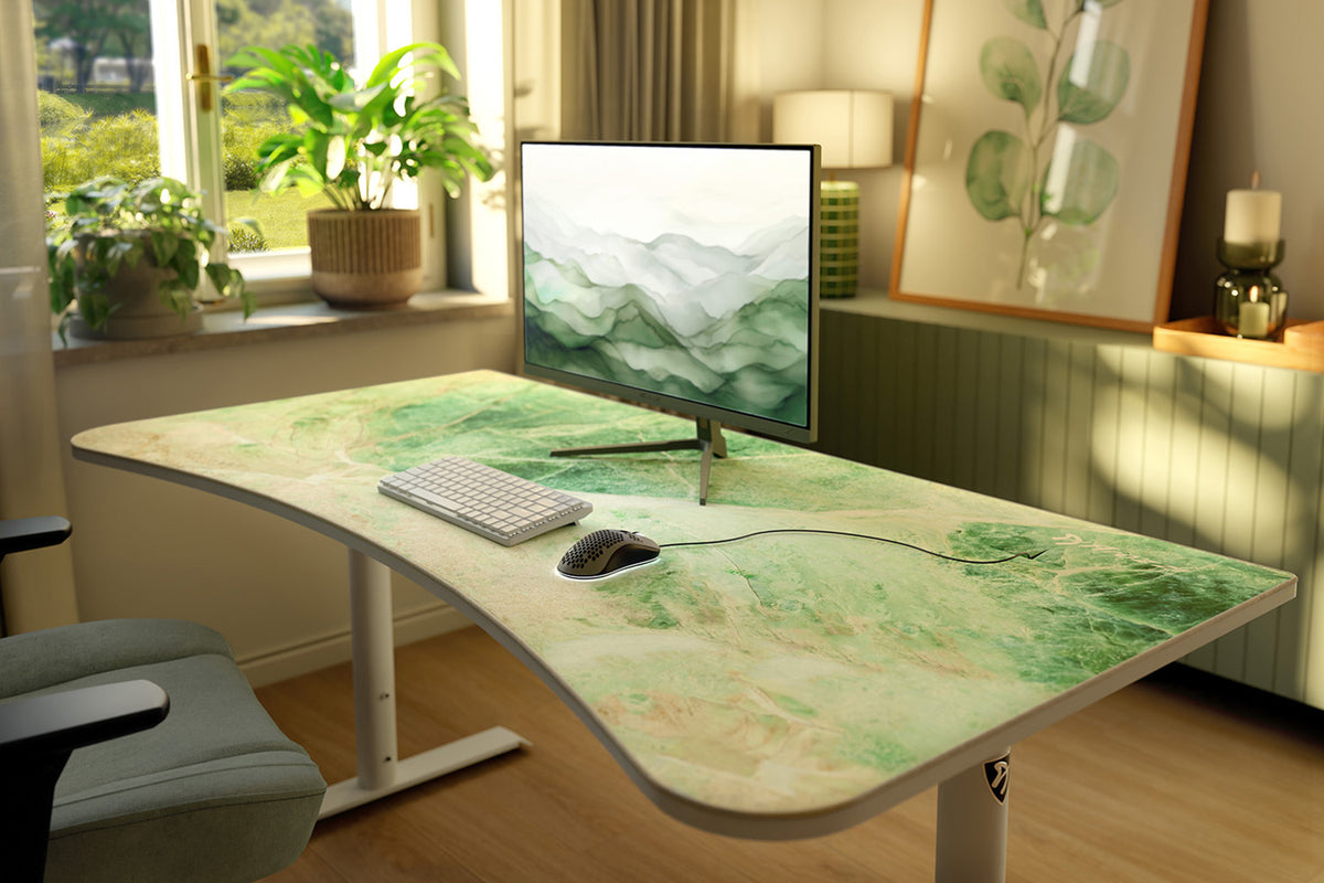 Curved desk with leafy design, monitor, keyboard, mouse, and plants by a sunny window.