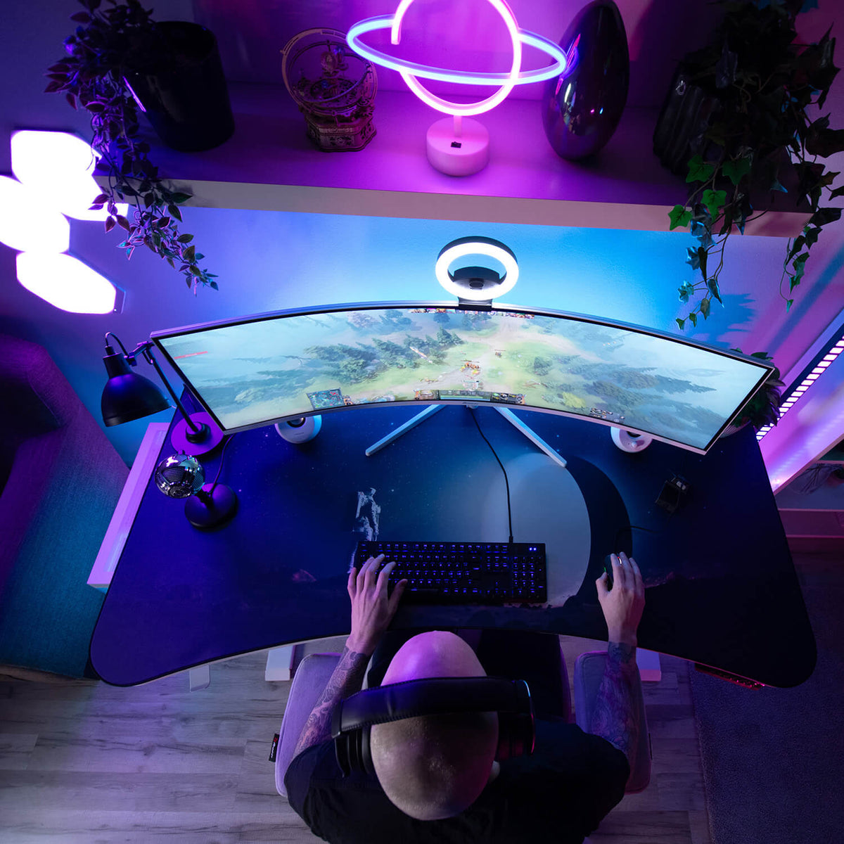 Person gaming at a curved monitor with purple neon lights and plants on the desk.