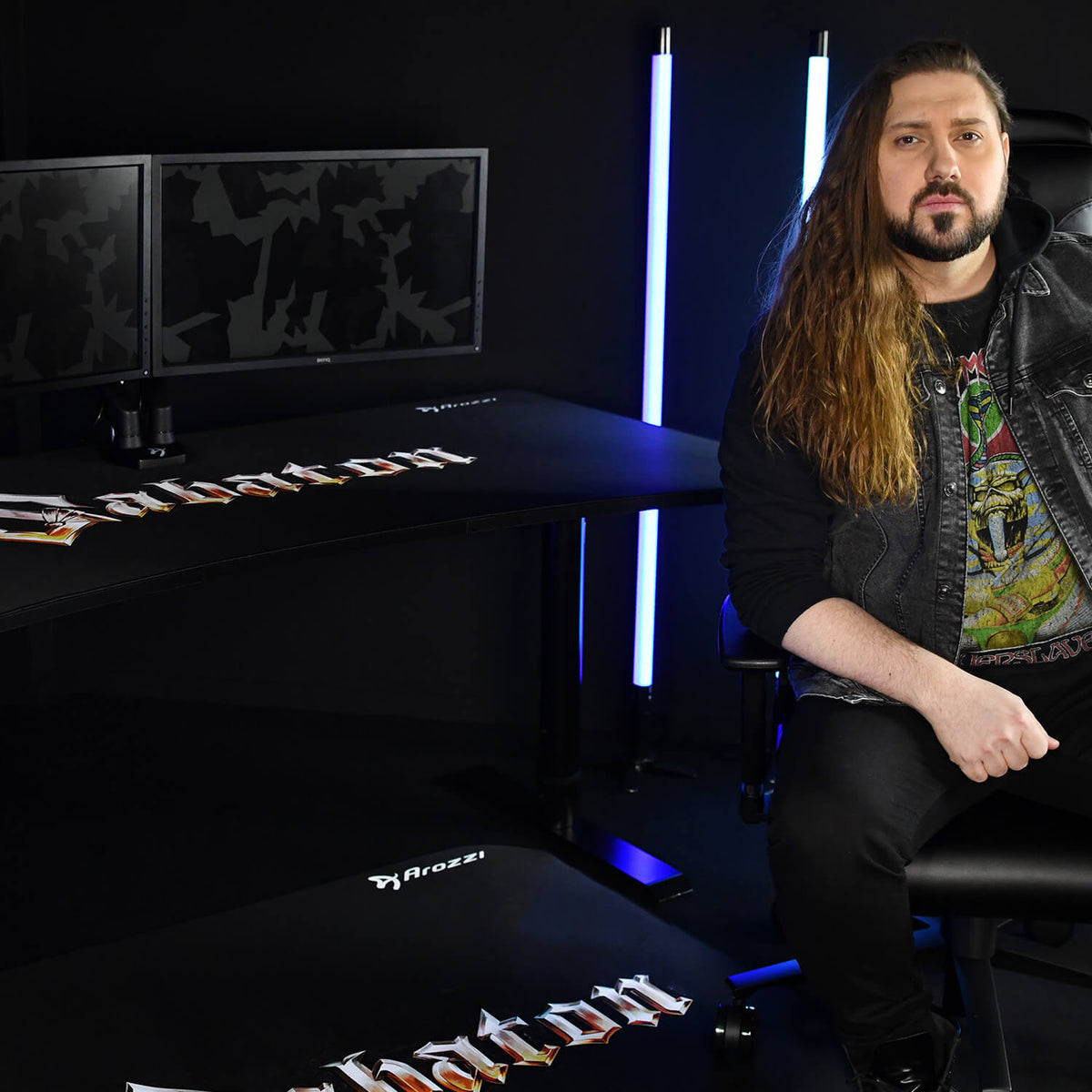 Man with long hair sits at Arozzi Arena Sabaton desk, neon blue lights glow behind monitors.