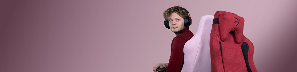 Person with curly hair wearing headphones sits in a gaming chair against a pink background.