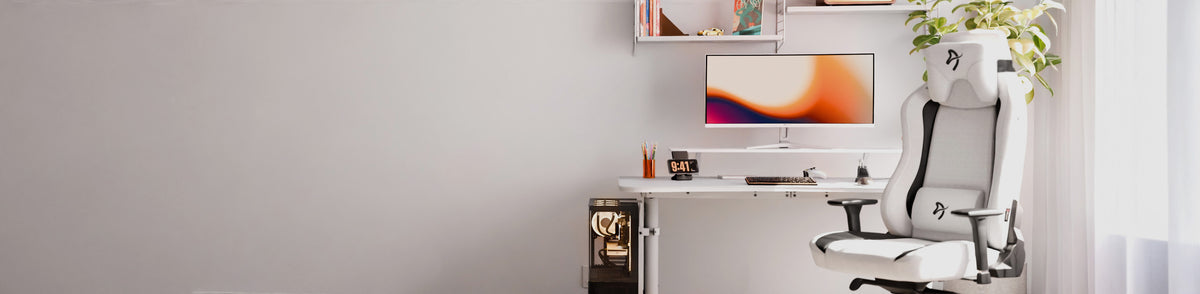 White office chair and desk with monitor, keyboard, books, clock, and plant by a window.