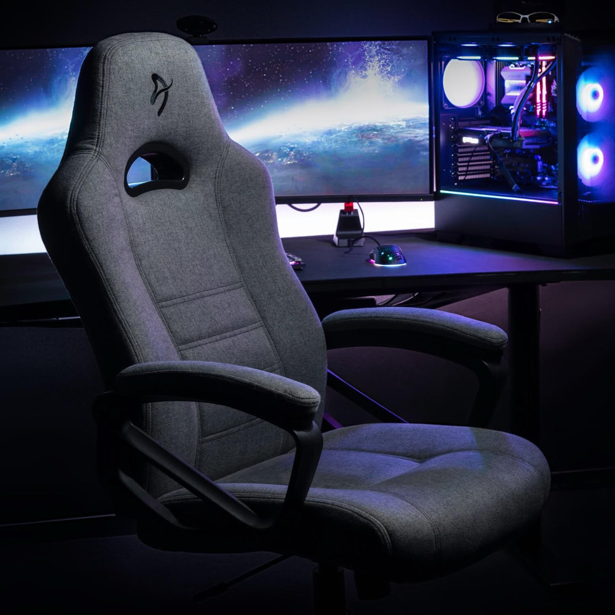 Gray gaming chair in front of a desk with monitors and a glowing PC in the background.