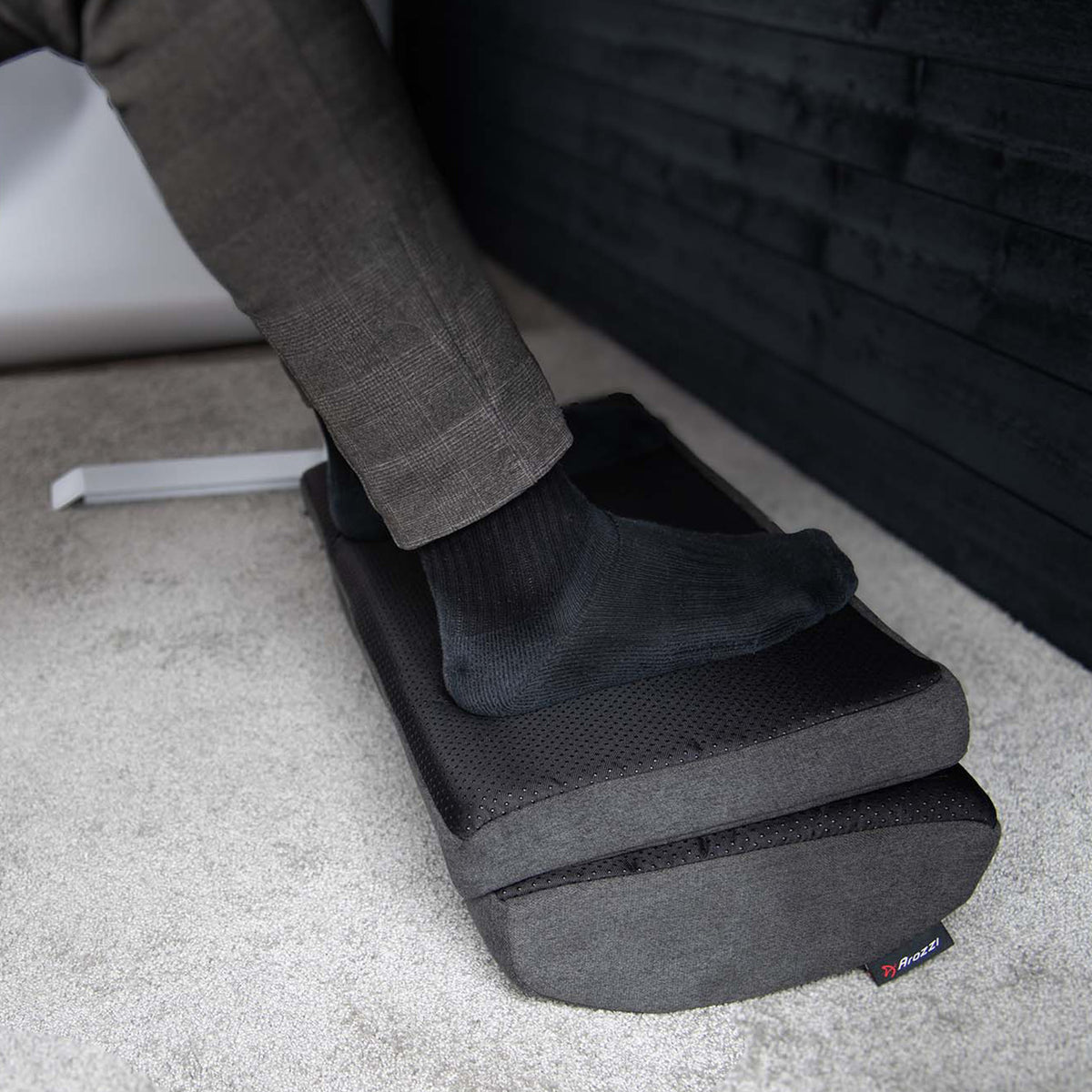 Arozzi Europe Footrest supports a socked foot under a desk on a carpeted floor.