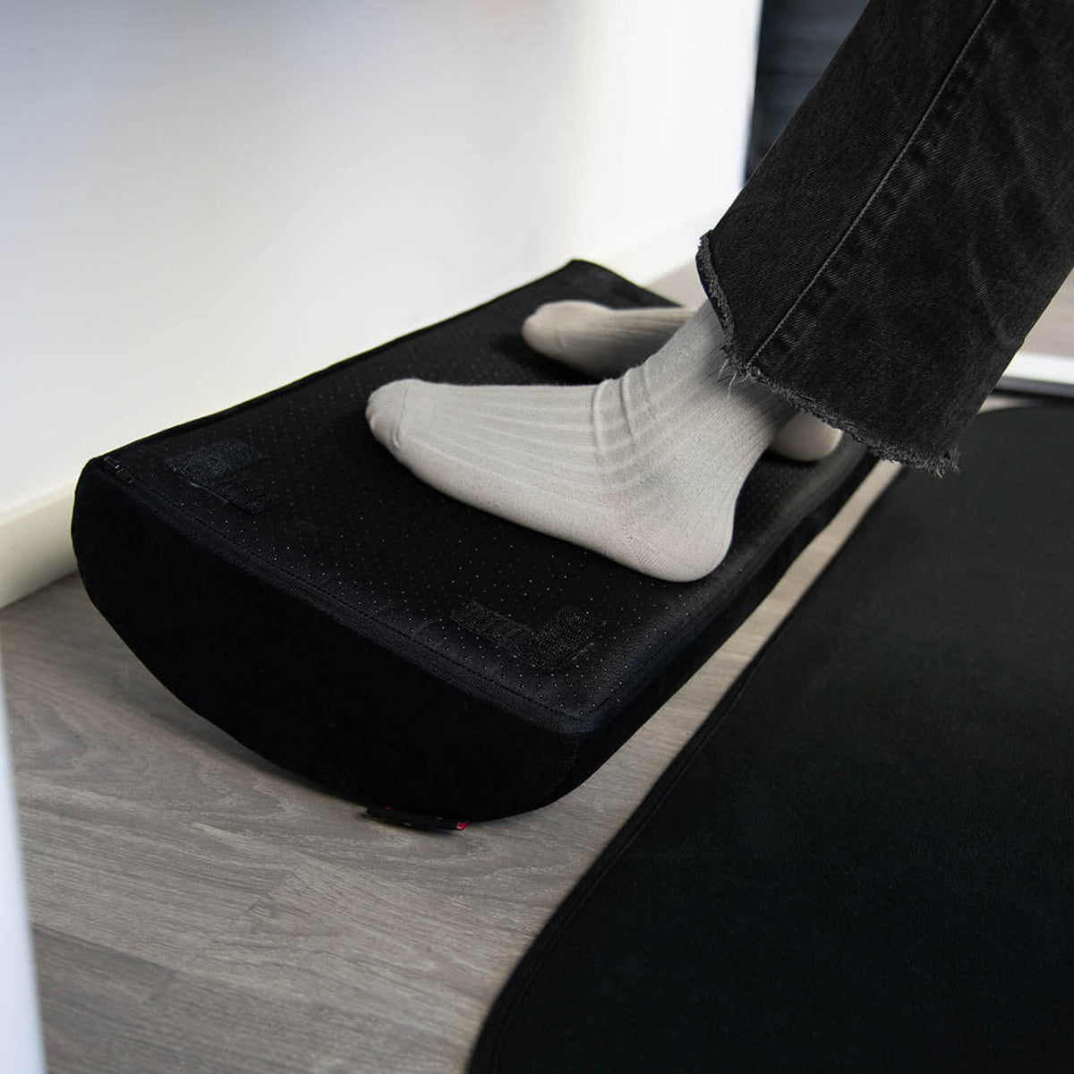 Person wearing socks rests feet on Arozzi Europe Footrest cushion on a wooden floor.