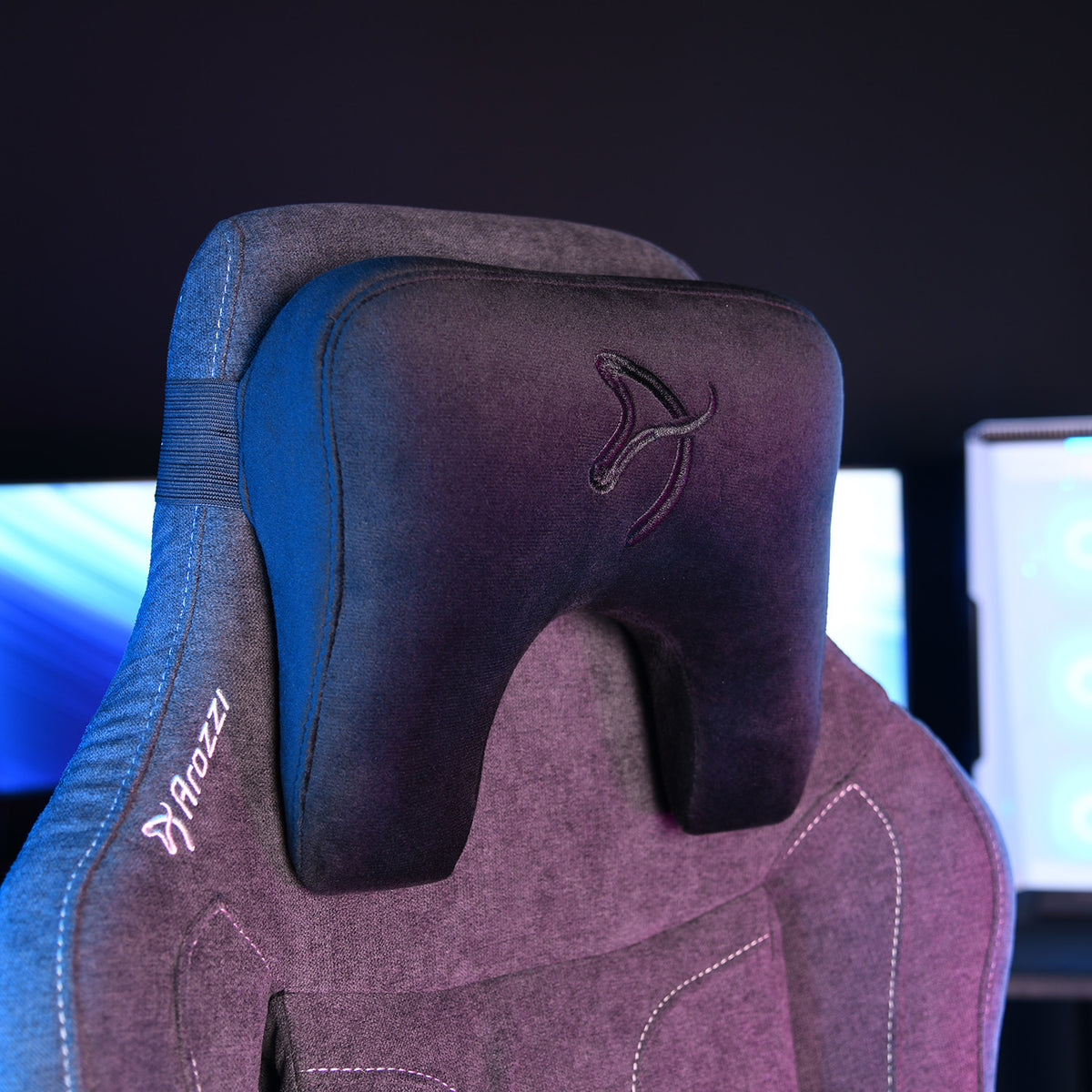 Arozzi Europe Neck Support Pillow Velvet Black on a gaming chair, screens glowing behind.