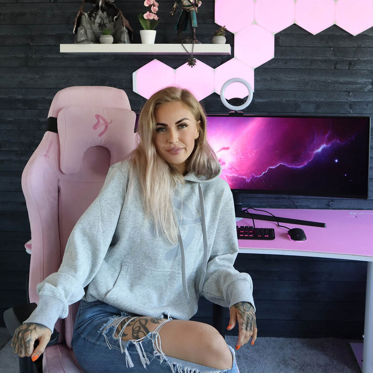 Blonde woman smiles at camera, seated on Arozzi Europe Vernazza Supersoft in pink gaming setup.