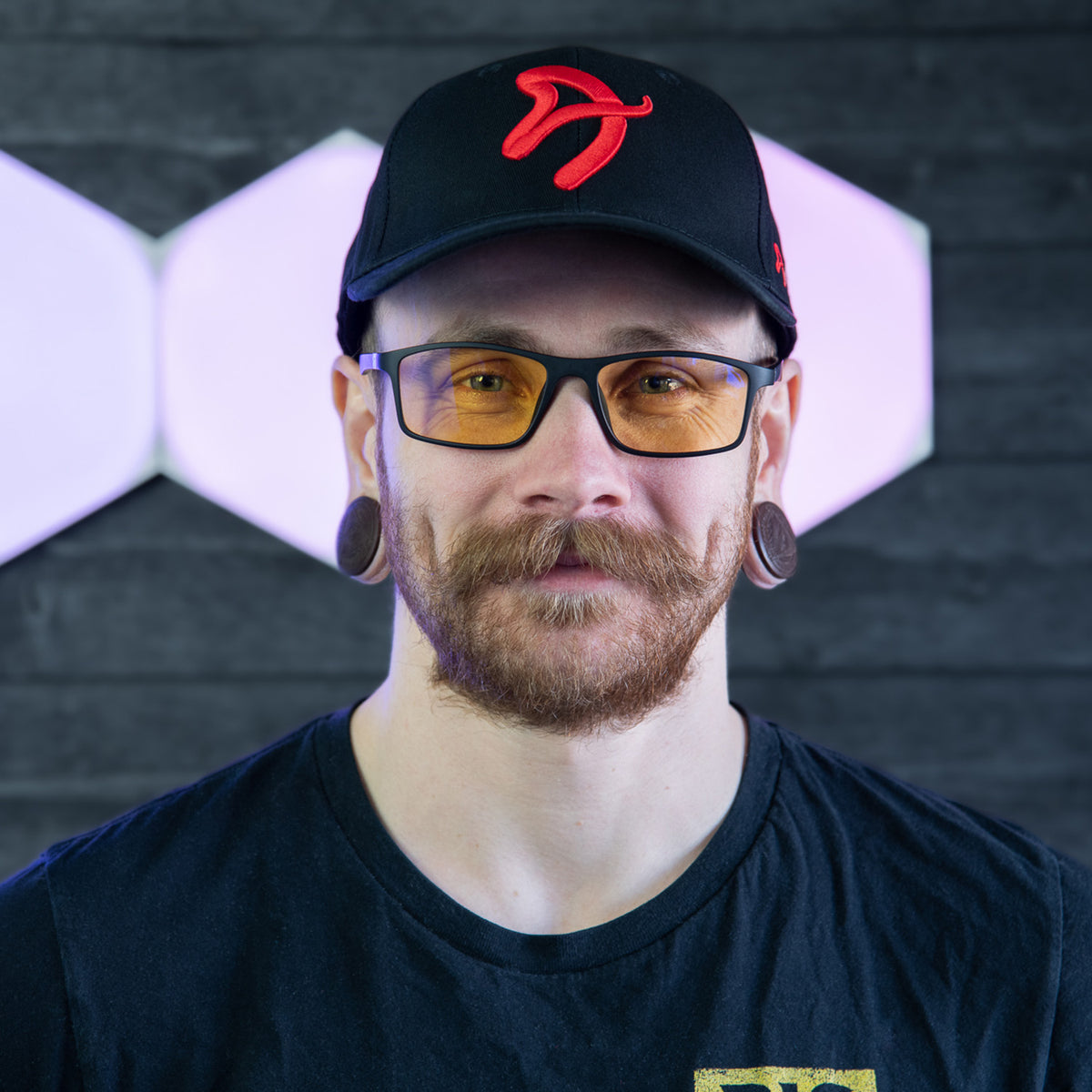 Man with glasses, mustache, red-cap, wearing Arozzi Europe Visione VX-200 by a lit wall.
