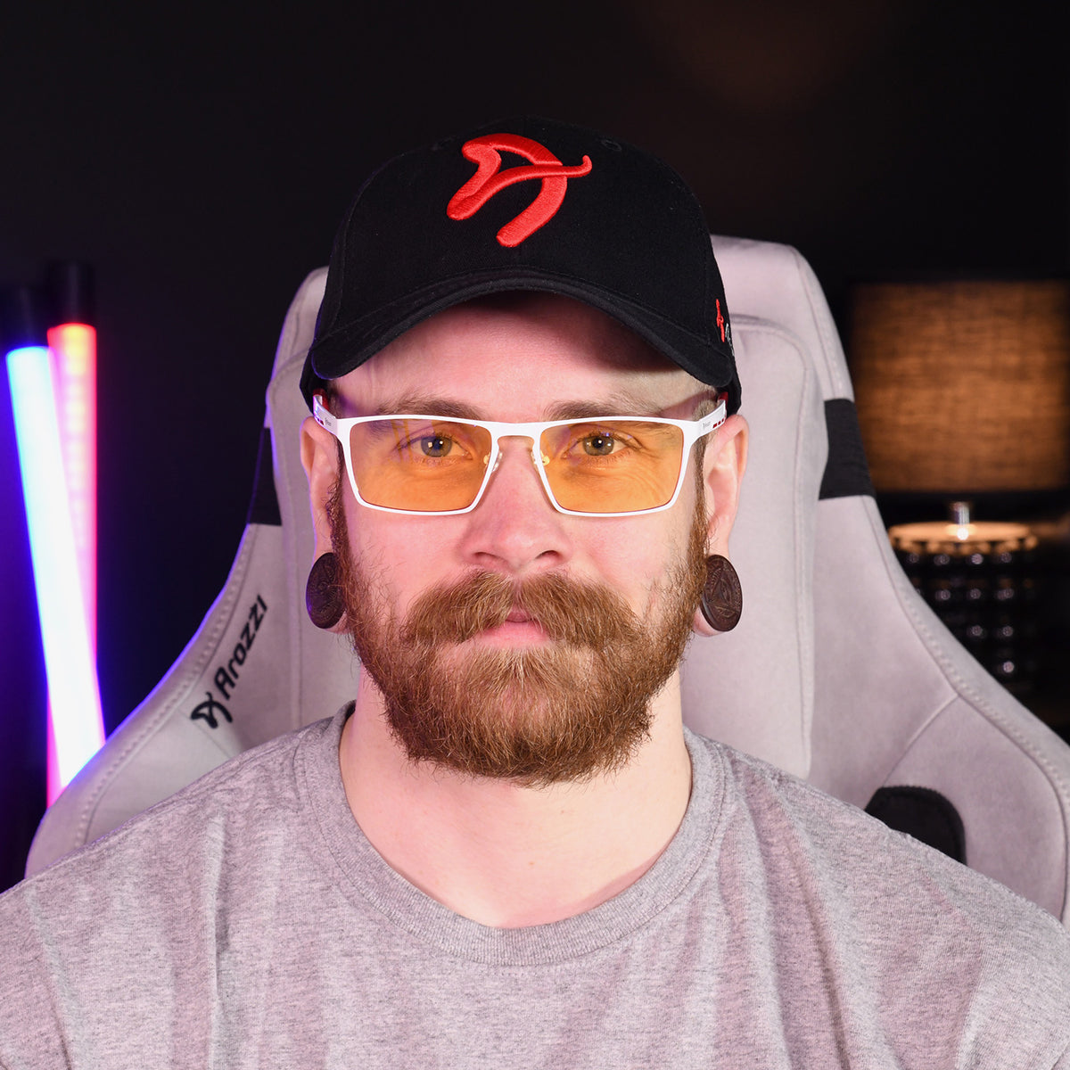 Man on gaming chair wearing Arozzi Visione VX-800 blue light glasses, cap & grey shirt.