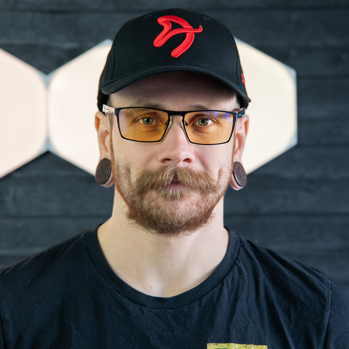 Man with Visione VX-800 blue light glasses by Arozzi Europe, cap, mustache, gauges, dark wall.