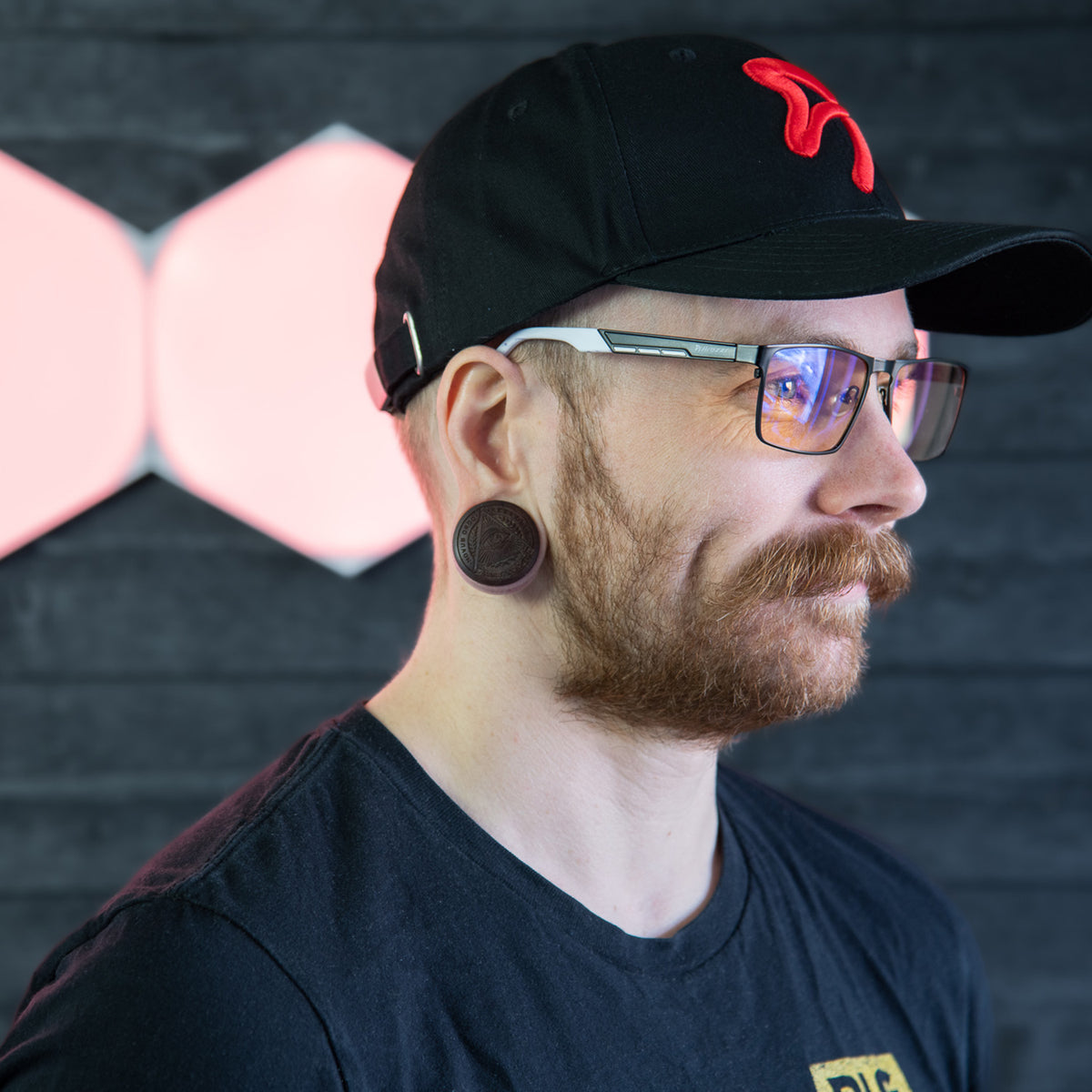 Man with Visione VX-800 glasses by Arozzi Europe, black cap, ear gauges, mustache, dark wall.