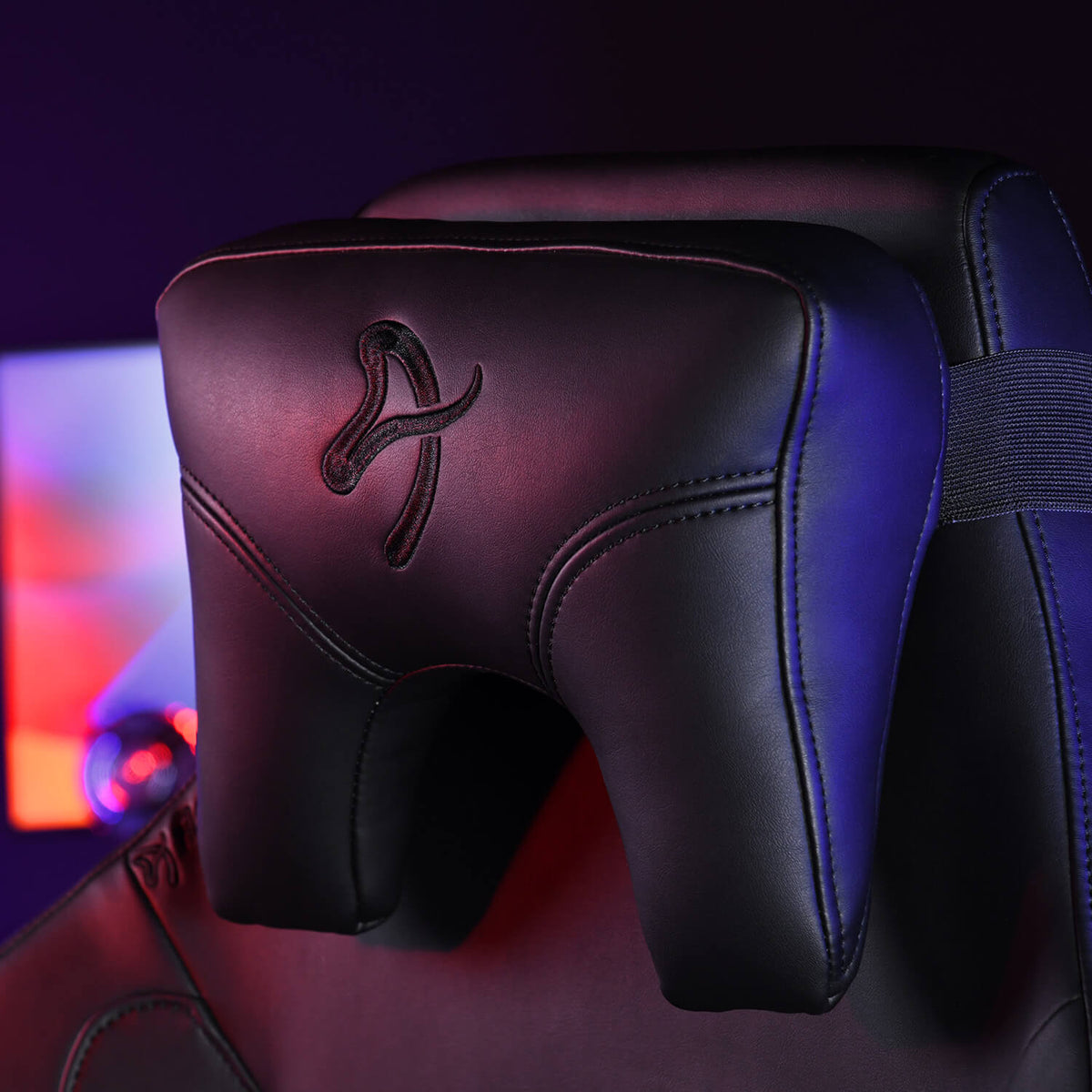 Black gaming chair headrest with stitched logo, lit by blue and pink lights.