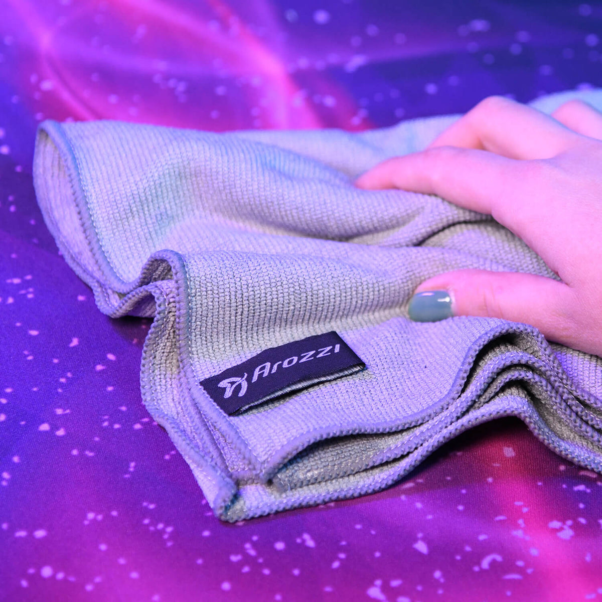 Hand holding gray cloth with Arozzi label on a purple, starry-patterned surface.