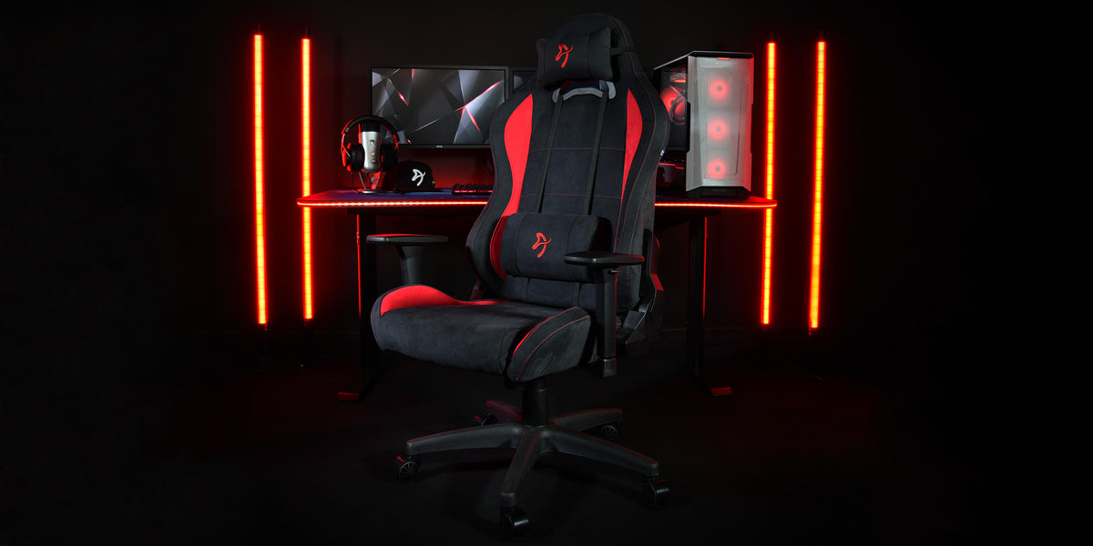 Black and red gaming chair in front of a desk with PC, monitors, headset, and red lights.