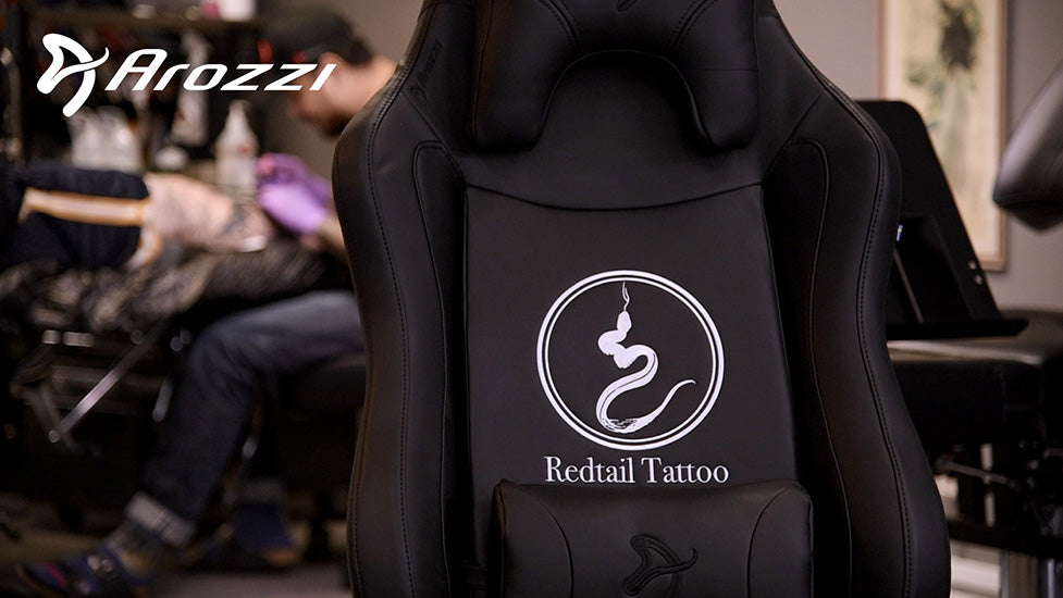 Black chair with Redtail Tattoo and snake logo; tattoo artist blurred in background.